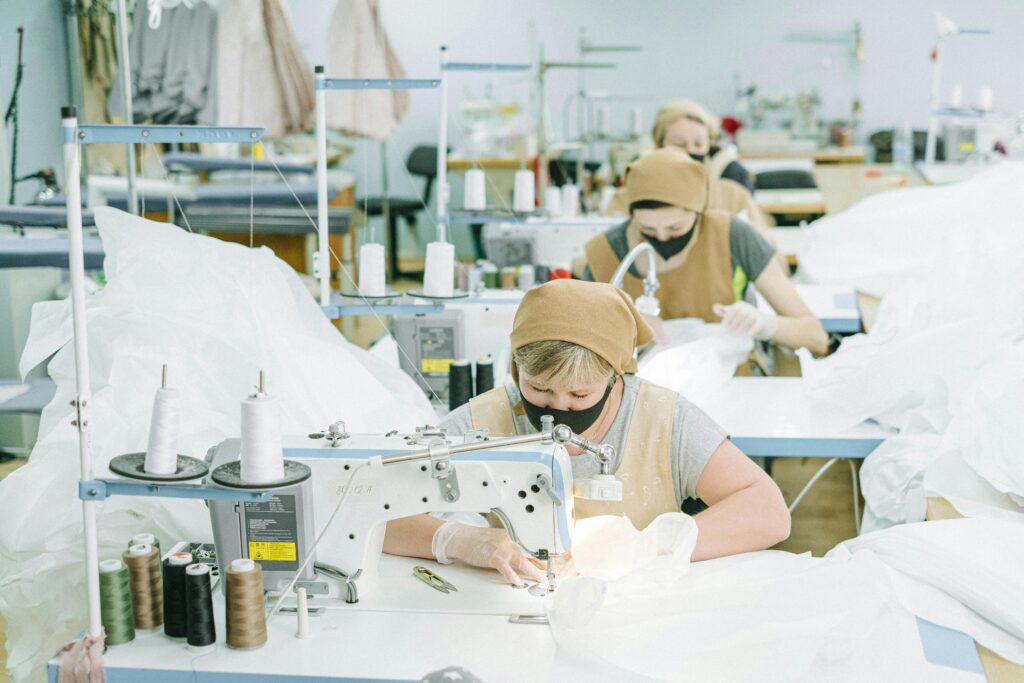 About Women working on sewing machines in an industrial textile workshop, wearing protective masks and aprons.