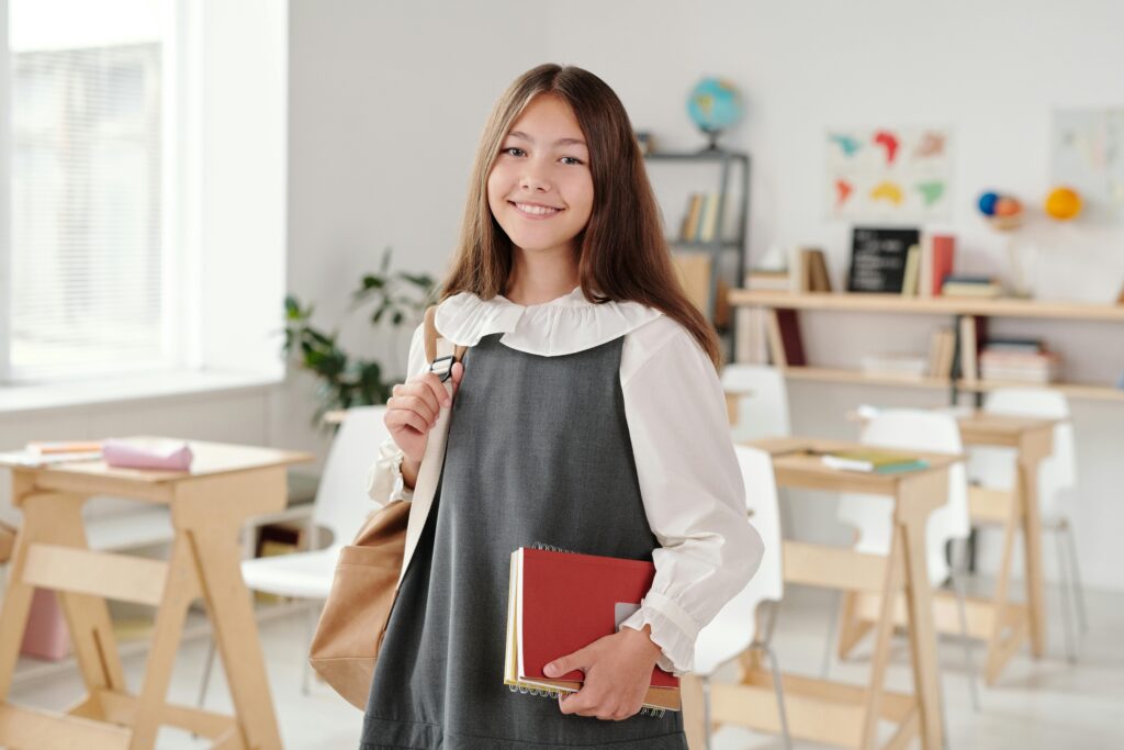 pexels photo 5211472 5211472 Smiling teenage girl in a school uniform holding notebooks in a bright classroom.